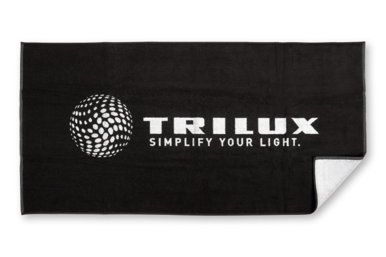 Duschtuch hingelegt mit einer umgeknickten Ecke, Schwarz mit weißem TRILUX Logo mittig groß, Ecke zeigt andere weiße Seite des Tuchs