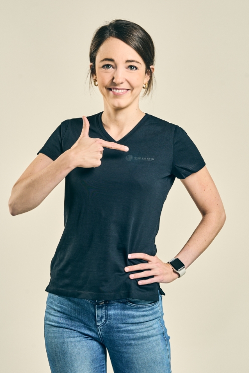 Weibliches Model trägt eine Jeanshose und ein schwarzes T-Shirt mit V-Ausschnitt, und zeigt mit dem Finger auf das weiße TRILUX Logo auf der linken Brust