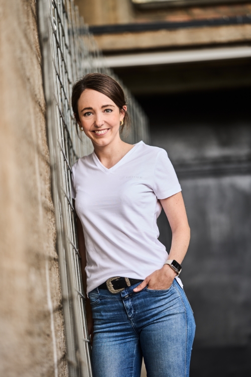 Weibliches Model lehnt an Wand, trägt Jeanshose und ein  Weißes T-Shirt mit V-Ausschnitt, weißes TRILUX auf linker Brust
