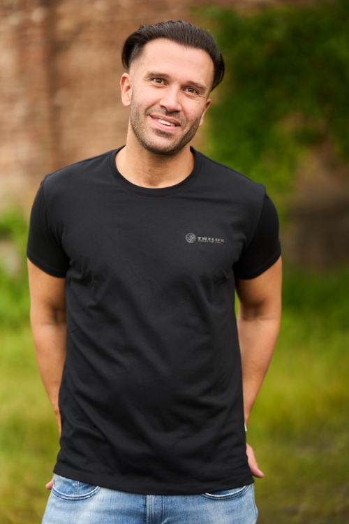 Männliches Model auf buntem Hintergrund trägt Jeanshose und schwarzes T-Shirt mit weißem TRILUX Logo auf linker Brust