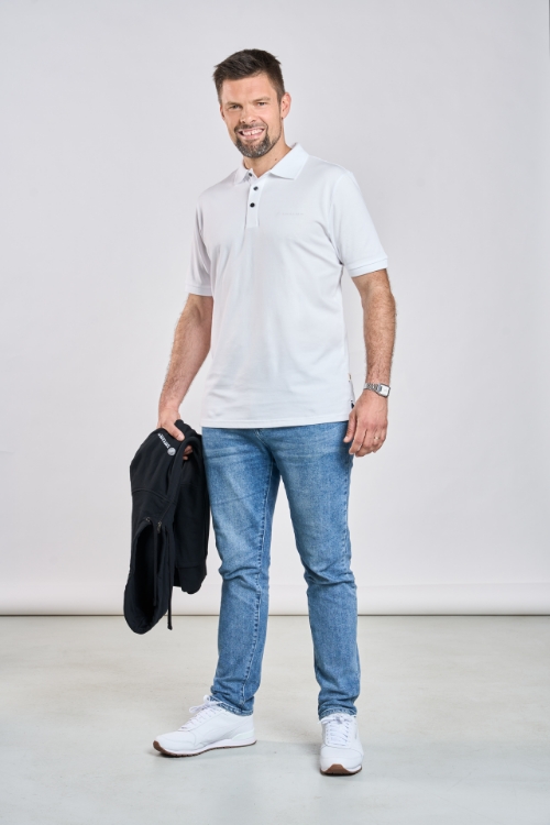 Männliches Model trägt Jeanshose, einen schwarzen Hoody in der Hand und ein Weißes Poloshirt mit drei schwarzen Knöpfen, weißes TRILUX Logo auf der linken Brust