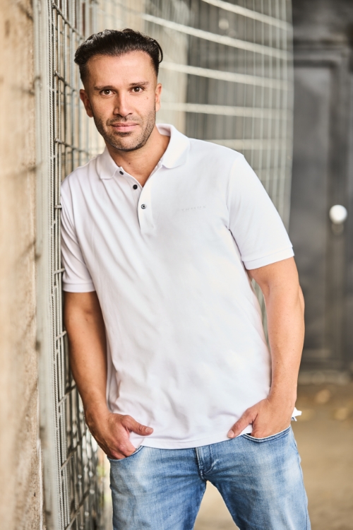 Männliches Model lehnt an einer Wand mit buntem Hintergrund trägt Jeanshose, einen schwarzen Hoody in der Hand und ein Weißes Poloshirt mit drei schwarzen Knöpfen, weißes TRILUX Logo auf der linken Brust